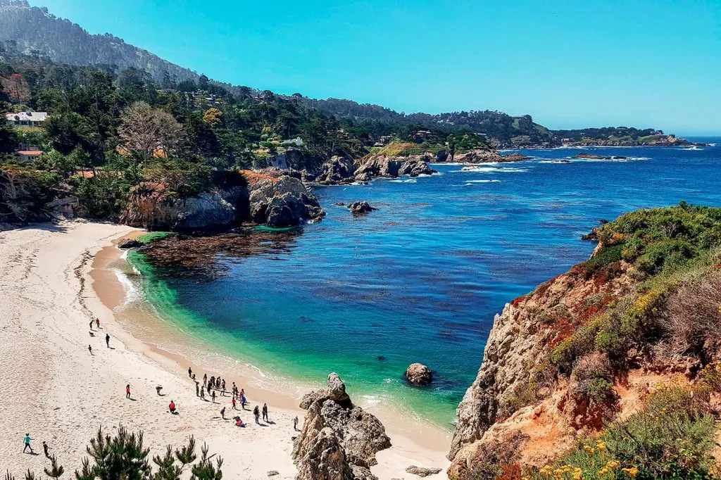 Beachgoers walking along a white sand beach on the rocky coastline of Carmel-by-the-Sea, California.